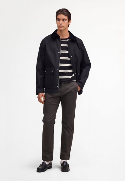 Homme portant une veste noire par-dessus une chemise rayée noir et blanc, avec un pantalon marron foncé et des mocassins noirs, debout la main dans la poche.