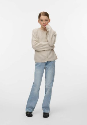 Jauna mergina stovinti su ranka prie smakro, dėvinti smėlio spalvos užsegamą megztinį, šviesiai mėlynas džinsus ir juodus batus, fone – paprasta balta siena.