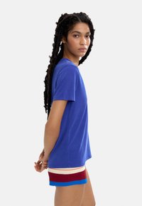 Niebieska krótko rękawowa koszulka z miękkiego materiału, o luźnym kroju. Modelka nosi kolorowe, prążkowane szorty w czerwono-biało-niebieskie paski.