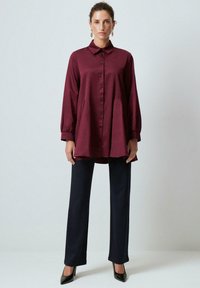 Woman standing, wearing a long burgundy button-up tunic, black straight-leg pants, black heels, and gold drop earrings against a plain wall.