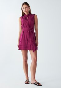 Trumpas rankovėmis, prigludęs suknelė tamsiai purpurnio spalvos su apykakle, sagčių priekyje ir derančia diržu. Juosmenyje yra horizontalių raukšlių.