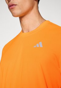 Πορτοκαλί αθλητική μπλούζα με κοντά μανίκια, στρογγυλή λαιμόκοψη, από λείο ύφασμα, με γκρι λογότυπο τριών ρίγων στο στήθος.