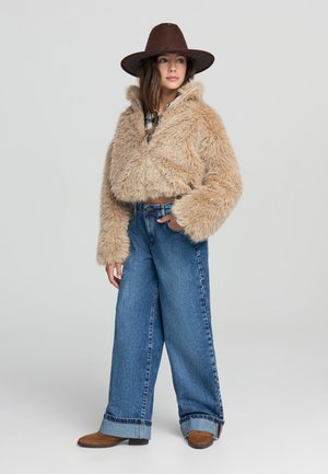 Donna con un cappello marrone a tesa larga, giacca corta in pelliccia sintetica color beige, jeans azzurri a gamba larga con risvolti e stivali in suede marroni, in piedi su un pavimento grigio.