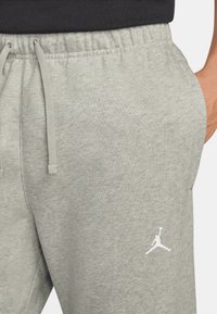 Pantalon de survêtement gris clair avec taille élastique et cordon de serrage, présentant une texture lisse et un logo Jumpman blanc sur la cuisse gauche.