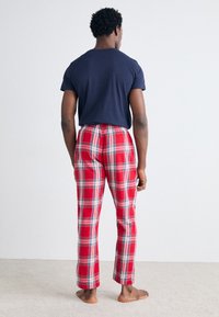 Røde ternede pyjamasbukser med blød tekstur og elastisk linning, kombineret med en ensfarvet mørkeblå t-shirt. Barfodet stående på et lyst tæppe.