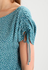 Turquoise top met witte polka dots, voorzien van geplooide pofmouwen en verstelbare linten. Gemaakt van zachte stof met een gestructureerd uiterlijk.