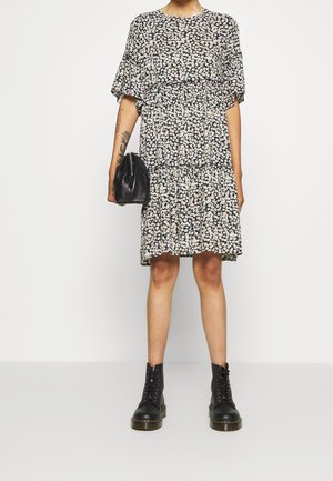 Person trägt ein schwarz-weißes Blumenkleid, hält eine schwarze Handtasche und trägt schwarze Schnür-Kampfboots, steht vor einem schlichten Hintergrund.