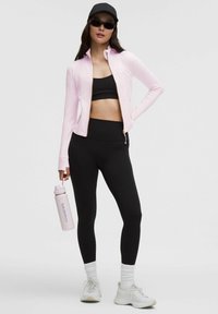 Jasnoróżowa kurtka sportowa na zamek, czarny top krótki, czarne legginsy z wysokim stanem, szare buty biegowe, białe skarpetki sięgające kostek oraz różowa butelka na wodę.