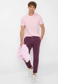 T-shirt rosa a maniche corte, pantaloni slim-fit bordeaux e sneaker bianche, che tiene in mano una giacca rosa chiaro. Design semplice, colori classici.