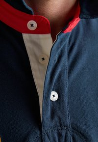 Polo bleu foncé avec un col rouge, une patte de boutonnage blanche, et trois boutons blancs, partiellement déboutonnés au niveau de l'encolure.