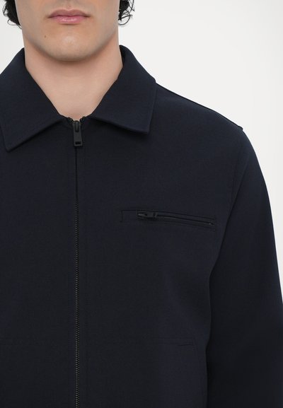 Mornarsko modra jakna z zadrgo, ki ima ovratnik, žep na prsih z zadrgo ter gladko teksturo blaga. Čista zasnova z minimalnimi detajli.