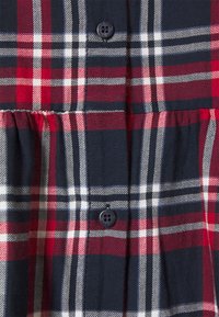 Tessuto a quadri rosso e blu navy con linee verticali e orizzontali, dotato di un dettaglio a pulsante nero al centro. La texture appare morbida e lavorata a trama.