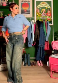 Kvinde iført en kort blå top og broderede jeans med vide ben står i et rum med farverige tøj hængende og par sko nedenunder.