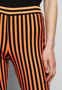 Svart och orange randiga leggings med en elastisk midja; tyget verkar slätt med en nära passform. Handen vilar på sidan.