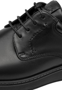 Gros plan d’une chaussure en cuir noire avec lacets, coutures visibles et col rembourré marqué sur la partie supérieure.