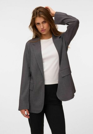 Jeune femme aux longs cheveux bruns, portant un blazer gris, une chemise blanche et un pantalon noir, posant avec une main sur la tête.