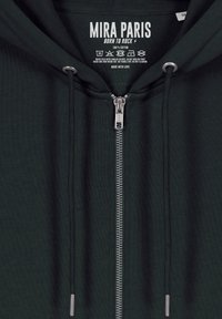 Felpa con zip in cotone verde scuro, dotata di zip metallica, cordini e un'etichetta con la scritta "MIRA PARIS - BORN TO ROCK." Texture morbida, design minimale.