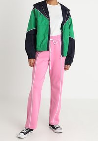 Person iført en grøn og sort vindjakke, hvid skjorte, lyserøde bukser med hvide side-striber og sorte sneakers, der står foran en hvid baggrund.