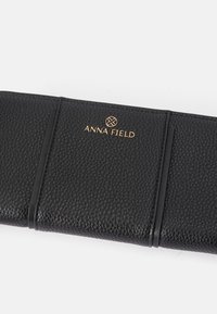 Juodas piniginė su akmeniniu tekstūru, su auksiniu iškilniu „ANNA FIELD“ logotipu ir plonu dizainu, turinti elastingą uždarymo detalę.