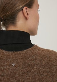 Brun tekstureret uldtrøje med vid halsudskæring, lagdelt over en sort ribbet turtleneck. Bemærkelsesværdig sølvfarvet logoaccent på ryggen.