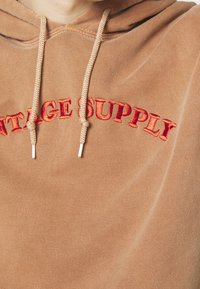 Brown hoodie with embroidered "VINTAGE SUPPLY" in red, featuring a drawstring hood and textured fabric.