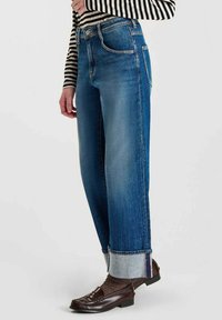 Jean en denim bleu clair avec une coupe décontractée, présentant des ourlets retroussés et un léger délavage. Associé à des chaussures marron foncé et un haut rayé.