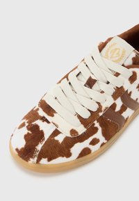 Sneaker con motivo di pelle di mucca marrone e bianco, con lacci beige, dettagli marroni e suola in gomma. La superficie texturizzata aggiunge un tocco di dettaglio.