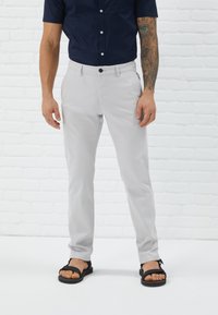 Pantalons gris clair à coupe ajustée, dotés d'une fermeture à bouton à l'avant et de poches, associés à des sandales noires, sur un fond de briques blanches.