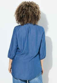 Camicia di jeans con maniche a tre quarti, orlo arrotondato e dettaglio plissettato sul retro. Colore azzurro chiaro, texture morbida, design casual.