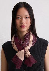 Foulard di seta in bordeaux e beige con motivi paisley. Indossato in modo sciolto attorno al collo, presenta bordi frangiati e una texture morbida.