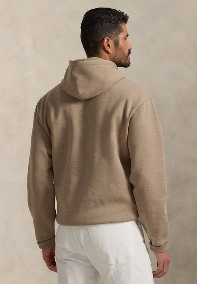 Felpa cappuccio color sabbia con una texture morbida, caratterizzata da una vestibilità comoda e polsini a coste, abbinata a pantaloni chiari.