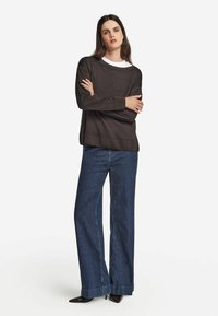 Kvinna står med armarna i kors, iklädd en brun långärmad tröja, vida blå jeans och svarta spetsiga skor mot en vit bakgrund.