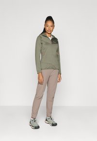 Chaqueta ligera de color verde oliva con cremallera y capucha, combinada con pantalones taper en tono beige y zapatos de senderismo grises, con tejido texturizado y acentos en naranja.