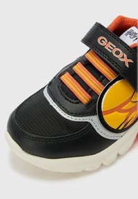 Zwarte sneaker met oranje en gele vlamontwerp, gemaakt van synthetisch leer, met klittenbandsluiting, textielstof en een witte rubberen zool.