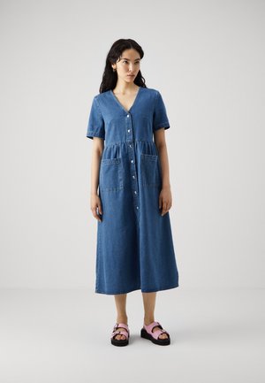 Blå denimkjole med korte ermer, V-hals, knappelukking foran og to store lommer. Kombinert med rosa sandaler med svarte såler.
