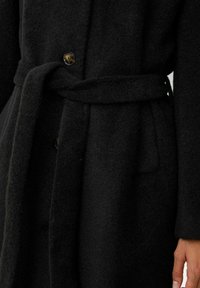 Zwarte wollen jas met een textuurafwerking, voorzien van een strikceintuur, grote knopen en zijzakken. Gekraagd ontwerp met lange mouwen.