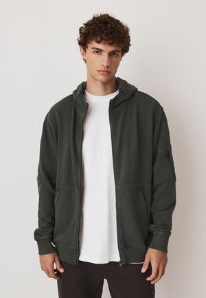 Jonge man met krullend haar die een donkergroene zip-up hoodie draagt over een wit T-shirt, staand tegen een effen lichte achtergrond.