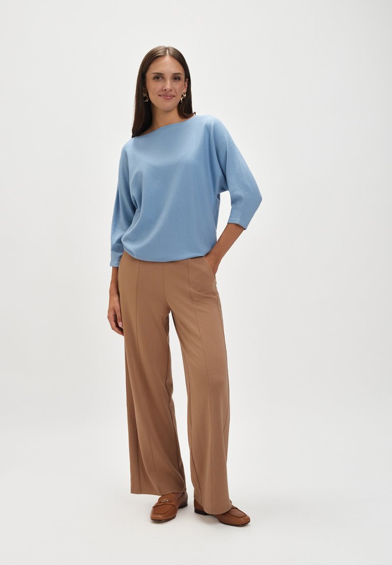 Jasnoniebieski sweter o szerokim dekolcie w połączeniu z beżowymi, zwiewnymi spodniami o szerokich nogawkach. Model nosi brązowe loafersy, podkreślając swobodny krój i miękką teksturę.