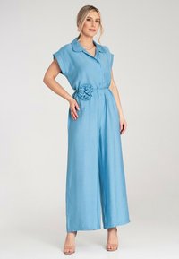 Niebieski lniany kombinezon z krótkimi rękawami, kołnierzykiem, zapinany na guziki z przodu oraz paskiem z detalem w postaci tekstylnego kwiatu. Szeroka nogawka.