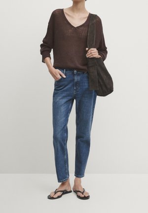 Person iført en løs brun V-hals sweater, blå jeans, sorte sandaler og bærende en mørkebrun ruskind skuldertaske.