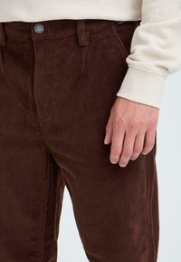 Brown corduroy pants with vertical ribbed texture, featuring a front pocket and a metal button. Partially visible hand resting on the fabric.