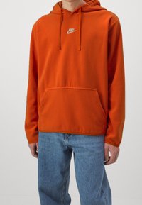 Man i ljus orange Nike-hoodie med framficka och blå jeans, stående mot en enkel ljus bakgrund.
