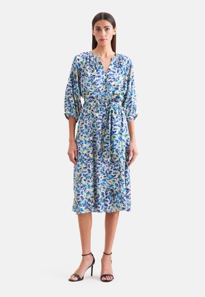 Femme debout portant une robe midi fleurie bleu et blanc avec des manches trois-quarts, une taille cintrée et des sandales à talons noirs.