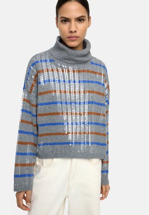 Kobieta ubrana w szary sweter z golfem z niebieskimi i brązowymi paskami oraz białymi cekinami, połączony z jasnobeżowymi spodniami.