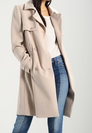 Femme portant un manteau beige à double boutonnage sur un haut blanc et un jean bleu bicolore, avec une main dans la poche du manteau.