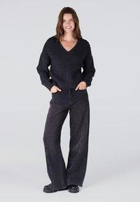 Zwarte, gebreide v-halstrui met een gestructureerd patroon, gecombineerd met hoge, wijde donkere jeans en zwarte plateau- laarzen.