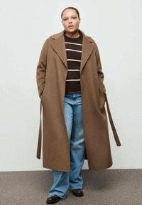 Brązowy wełniany płaszcz z szerokim kołnierzem, zestawiony z brązowym i białym swetrzem w paski oraz jasnoniebieskimi dzwonowymi dżinsami. Całość dopełniają ciemne buty.