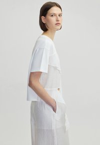 White layered top featuring short sleeves and a textured panel design, worn with a long, light-colored, pocketed skirt.