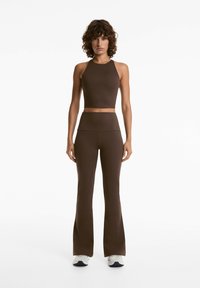 Barna bordázott pánt nélküli crop top, amelyhez illeszkedő, bővülő leggings tartozik; mindkettő sima textúrájú és testhezálló fazonú, fehér sportcipőkkel kiegészítve.