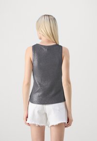 En blond kvinna sedd bakifrån, iförd en ärmlös glansig mörkgrå topp och vita fransade denimshorts mot en vit bakgrund.